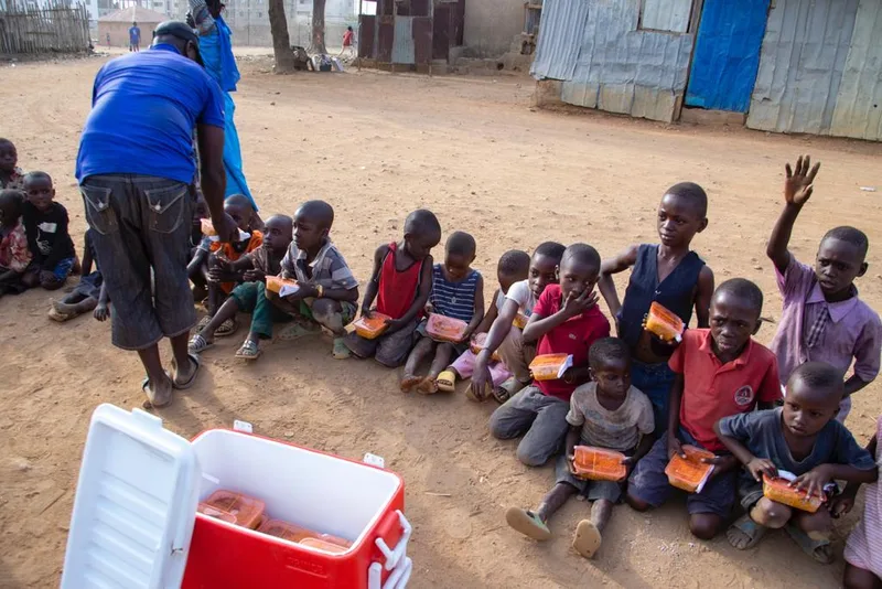 Children seated on the ground holding meal containers, beside a cool box.
